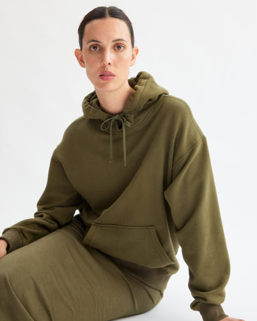A woman with dark hair pulled back wears the Arlo Mott Windsor Hooded Sweatshirt in Olive and a matching skirt, sitting against a plain white background while looking directly at the camera.
