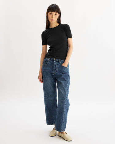 A woman with straight dark hair wears the Arlo Mott Bergen Short Sleeve Tee in Black, styled with loose blue jeans and cream-colored shoes, standing against a plain white background with one hand in her pocket.