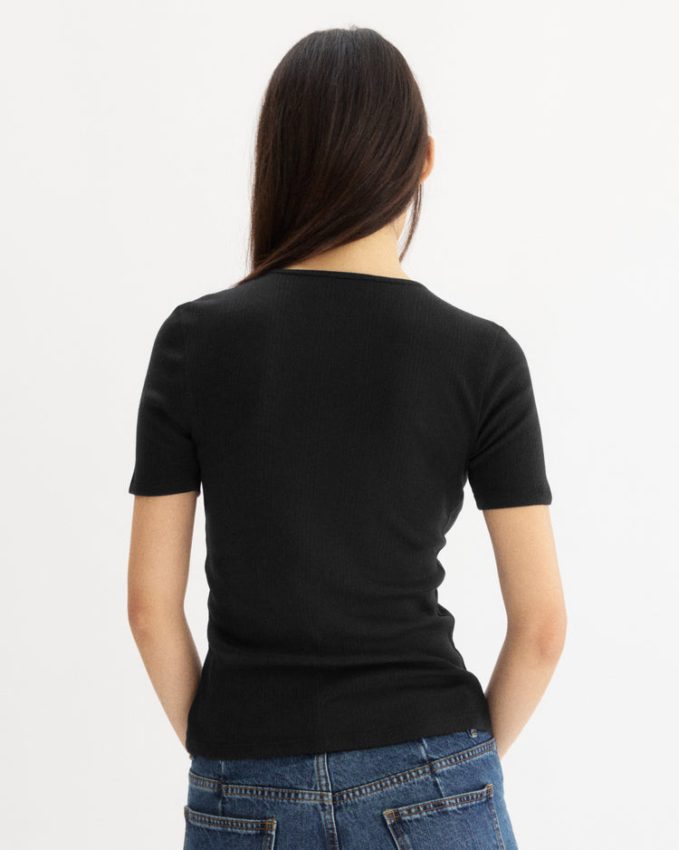 A woman with long brown hair faces away, wearing the Arlo Mott Bergen Short Sleeve Tee in Black and blue jeans against a plain white background.