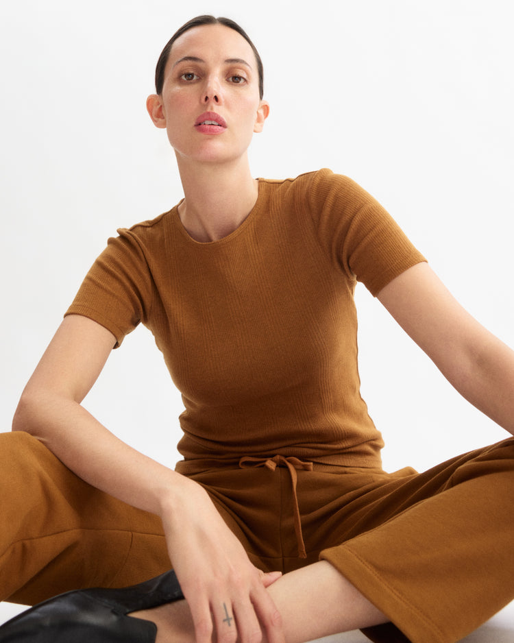 A woman with dark hair tied back, wearing the Arlo Mott Bergen Short Sleeve Tee in Cinnamon and matching pants, sits cross-legged on the floor against a plain white background. She has a neutral expression and a small cross tattoo on her finger.
