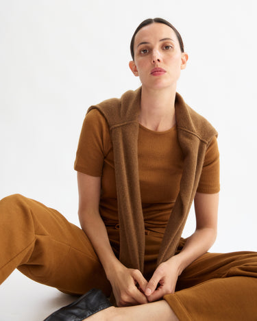 A woman sits on the floor against a white background, wearing the Arlo Mott Bergen Short Sleeve Tee in Cinnamon and matching slim fit pants, with a sweater draped over her shoulders. She looks directly at the camera with a neutral expression.