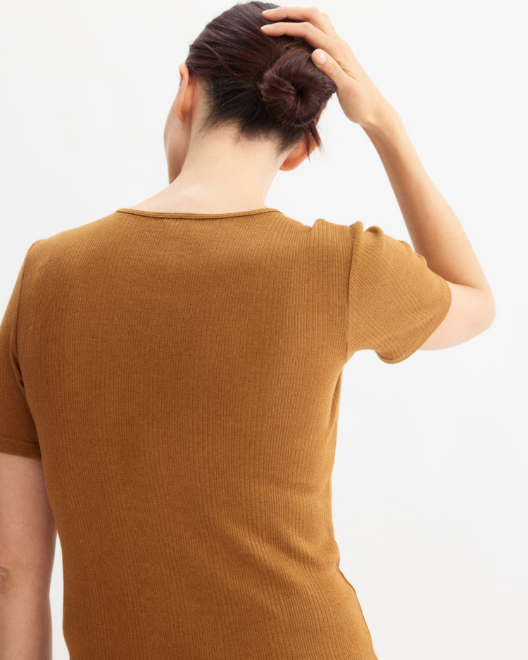 A person with dark hair in a bun faces away, wearing the Arlo Mott Bergen Short Sleeve Tee in Cinnamon and touching the back of their head against a plain light background.
