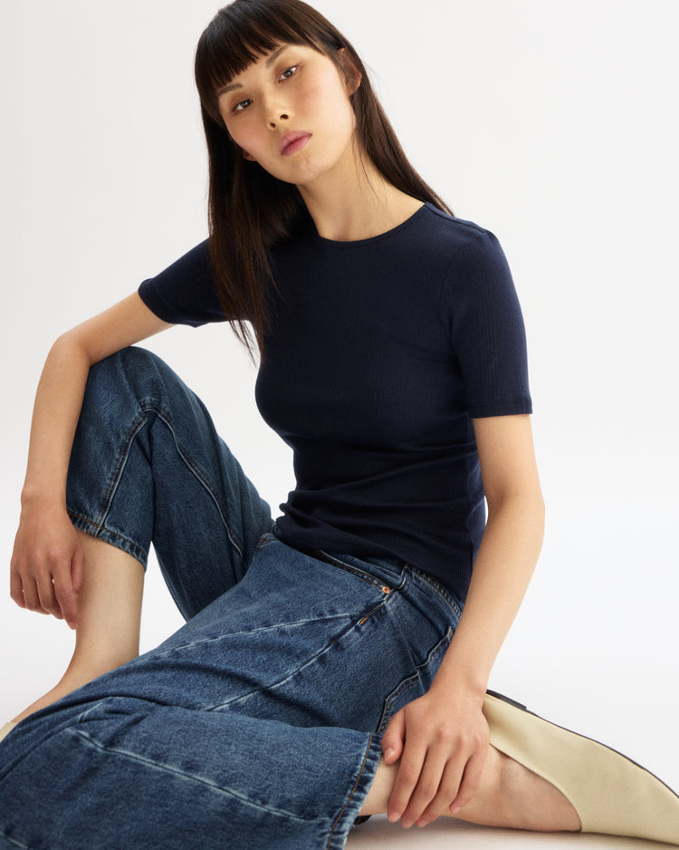 A woman with straight dark hair and bangs sits on the floor against a white background, wearing the Arlo Mott Bergen Short Sleeve Tee in Navy and wide-leg blue jeans, with one leg bent and the other extended.