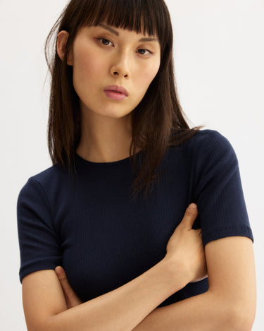 A woman with straight, dark hair and bangs stands against a plain background, arms folded and looking serious. She is wearing the Arlo Mott Bergen Short Sleeve Tee in Navy, a slim-fit ribbed style with short sleeves.