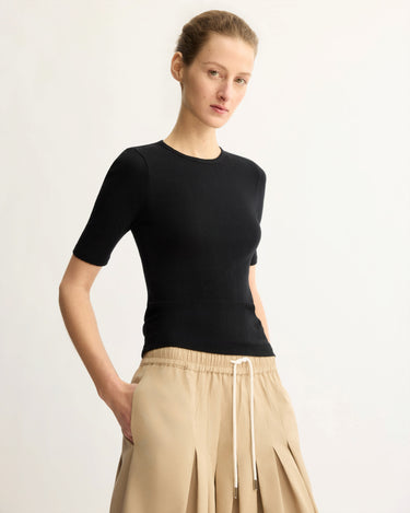 A person stands against a plain light background, wearing the Arlo Mott Quincy Slim Fit Tee in Black and loose beige pleated pants with a white drawstring, creating a slim silhouette while looking slightly to the side with hands in pockets.