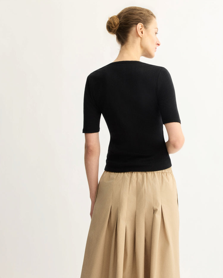 A woman with blonde hair in a bun, seen from behind, wears the Arlo Mott Quincy Slim Fit Tee in Black and a pleated beige skirt, showcasing her slim silhouette against a plain light background.