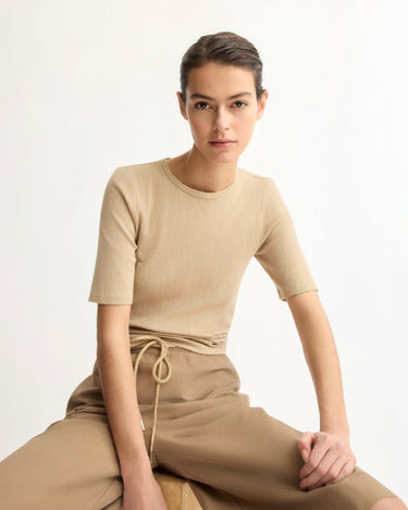 A woman with dark hair tied back wears the Arlo Mott Quincy Slim Fit Tee in Khaki, paired with matching pants. Seated on a wooden stool against a plain light background, she looks calmly at the camera in a relaxed pose.