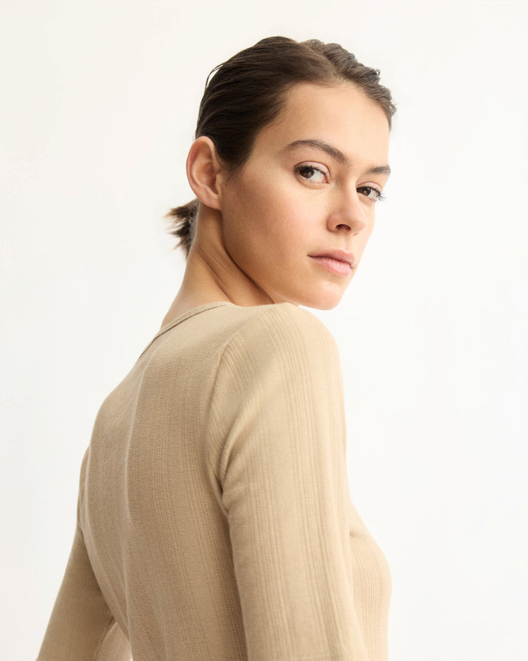 A woman with light skin and brown hair pulled back wears the Arlo Mott Quincy Slim Fit Tee in Khaki, posing with a glance over her shoulder toward the camera against a plain light background.