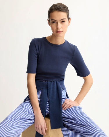 A woman sits on a wooden stool against a plain background, wearing the Arlo Mott Quincy Slim Fit Tee in Navy and blue-and-white striped pants, with a navy sweater tied at her waist as she looks confidently at the camera.