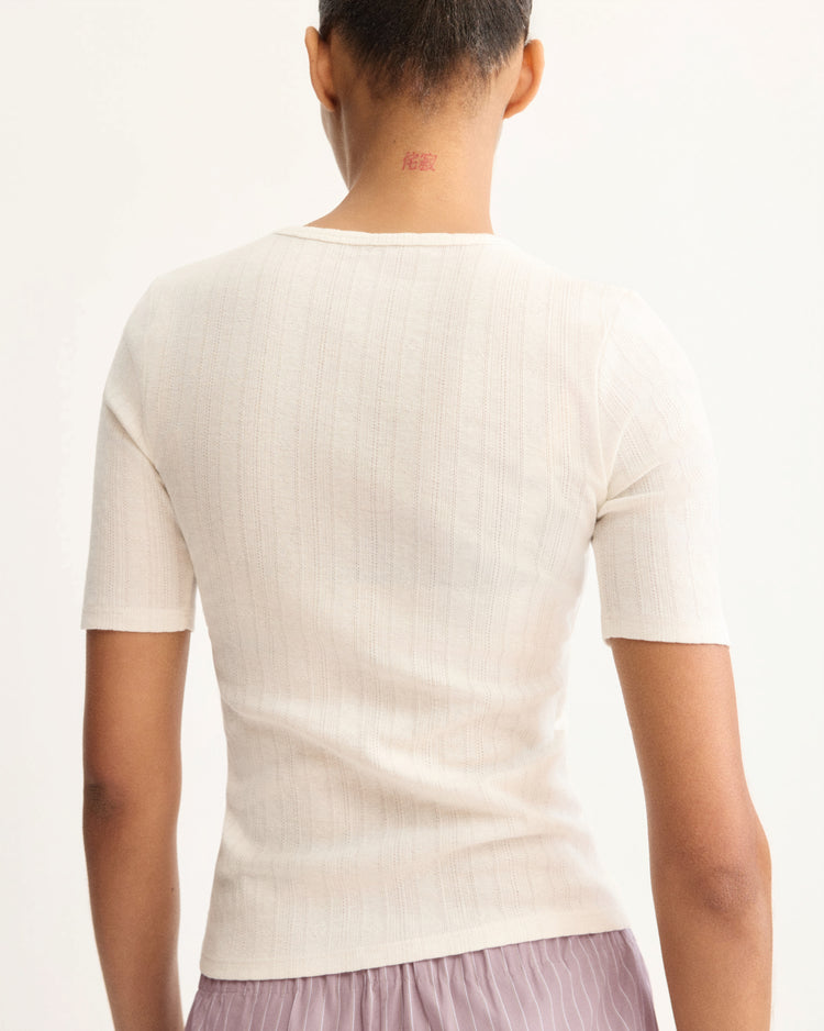 A person with short hair is seen from behind wearing the Arlo Mott Quincy Slim Fit Tee in White and a light pink skirt, with a small red tattoo on their neck accentuating the tee's slim silhouette.