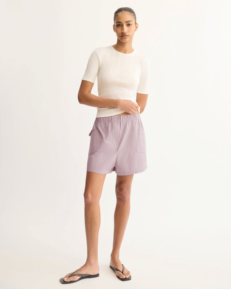 A person stands against a light background, wearing the Arlo Mott Quincy Slim Fit Tee in White, pink and white striped shorts with pockets, and black flip-flops. Their hair is pulled back and one hand rests in a pocket.