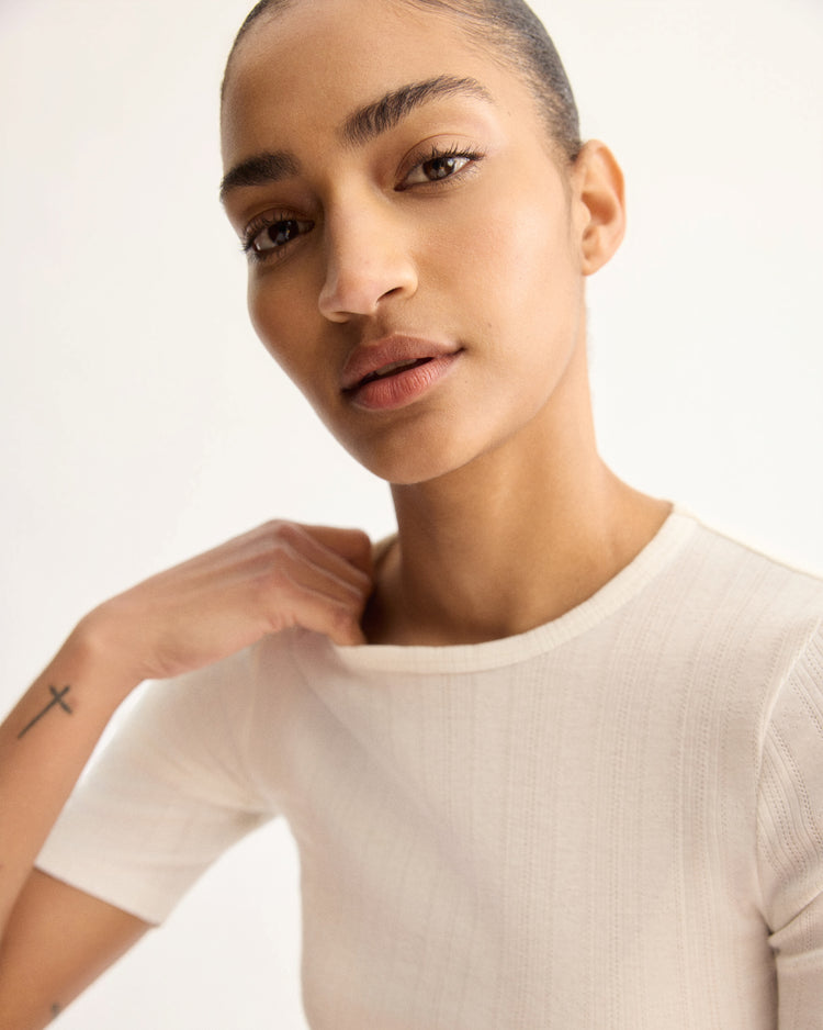 With smooth hair and natural makeup, a person gazes at the camera, gently holding the collar of the Arlo Mott Quincy Slim Fit Tee in White. A small cross tattoo on their forearm and a slim silhouette stand out against a plain light background.