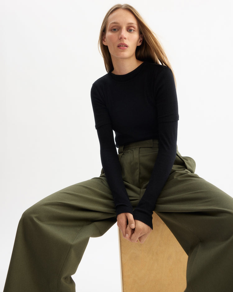 A woman with long blonde hair sits on a wooden block against a white background, wearing the Arlo Mott Clark Long Sleeve Tee in Black and high-waisted, wide-leg olive green pants for an elegant look as she gazes calmly past the camera.