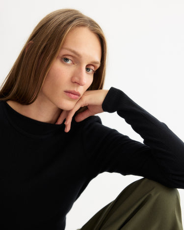 A person with long light brown hair wears the Arlo Mott Clark Long Sleeve Tee in Black and olive green pants, sitting with one arm on their knee and gazing thoughtfully at the camera against a plain white background.