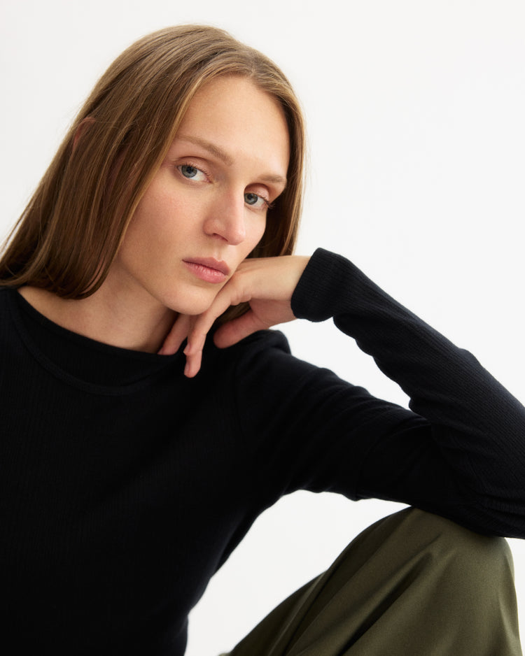 A person with long light brown hair wears the Arlo Mott Clark Long Sleeve Tee in Black and olive green pants, sitting with one arm on their knee and gazing thoughtfully at the camera against a plain white background.