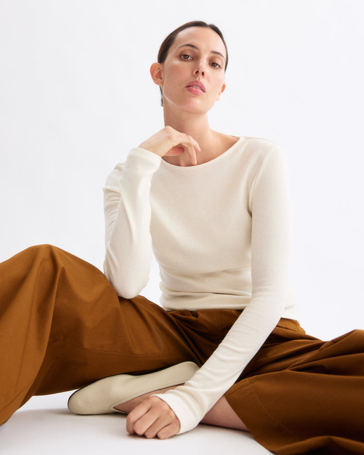 Wearing the Arlo Mott Clark Long Sleeve Tee in Chalk, a person sits on the floor with one leg bent and the other extended, creating an elegant silhouette against a plain white background.