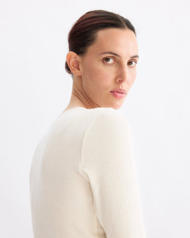 A woman with dark hair tied back glances over her shoulder at the camera, wearing the Arlo Mott Clark Long Sleeve Tee in Chalk, which highlights her elegant silhouette against a plain white background.