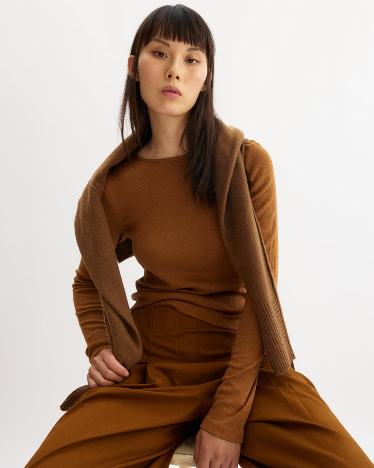 A woman with straight dark hair and bangs sits against a white background, wearing the Arlo Mott Clark Long Sleeve Tee in Cinnamon with wide-leg pants and a brown sweater draped over her shoulders for an elegant silhouette.