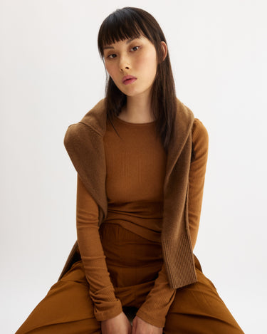 A woman with straight, dark hair and bangs sits on a stool against a white background. She wears the Arlo Mott Clark Long Sleeve Tee in Cinnamon over an Italian cotton T-shirt, paired with matching pants and a brown sweater draped over her shoulders.