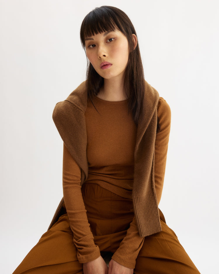 A woman with straight, dark hair and bangs sits on a stool against a white background. She wears the Arlo Mott Clark Long Sleeve Tee in Cinnamon over an Italian cotton T-shirt, paired with matching pants and a brown sweater draped over her shoulders.