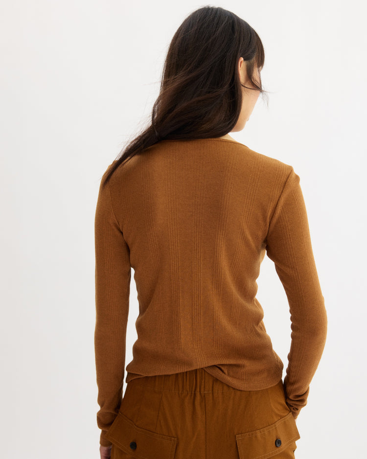 A person with long brown hair, wearing the Arlo Mott Clark Long Sleeve Tee in Cinnamon and brown pants, stands facing away from the camera against a plain white background.