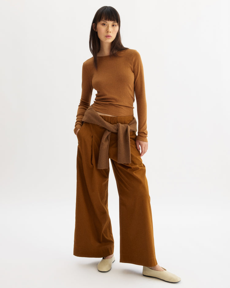 A woman with straight dark hair and a neutral expression stands against a white background wearing the Arlo Mott Clark Long Sleeve Tee in Cinnamon, wide-leg brown pants, a sweater tied at her waist, and cream-colored shoes.