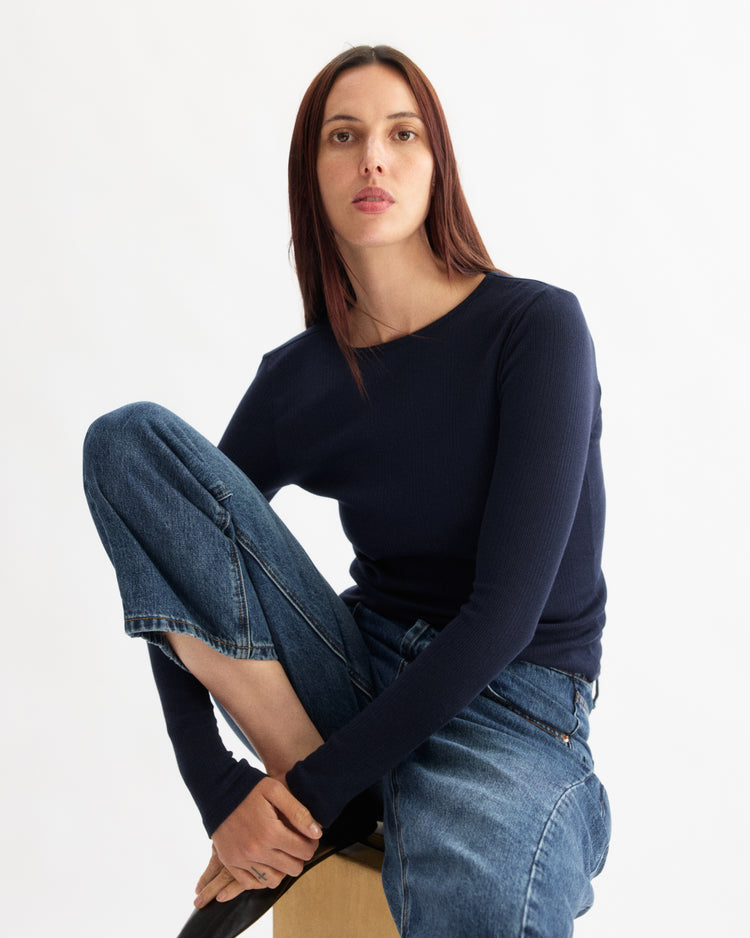 A woman with long brown hair wears the Arlo Mott Clark Long Sleeve Tee in Navy and blue jeans. She sits on a wooden block with one knee up, looking directly at the camera against a plain white background.