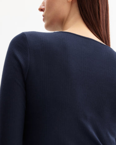 A close-up shows a woman with long brown hair wearing the Arlo Mott Clark Long Sleeve Tee in Navy, shown from the back and slightly to the side to highlight its elegant silhouette.