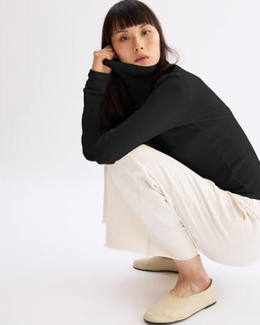 A woman with straight dark hair and bangs squats on the floor, wearing the Arlo Mott Clinton Long Sleeve Turtleneck in Black, cream wide-leg pants, and beige flats, with a neutral expression against a plain white background.