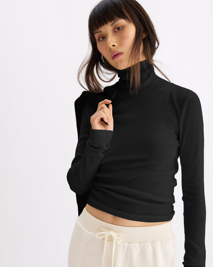 A woman with straight brown hair and bangs wears the Arlo Mott Clinton Long Sleeve Turtleneck in Black, paired with cream drawstring pants, standing against a light background and looking at the camera with a neutral expression.