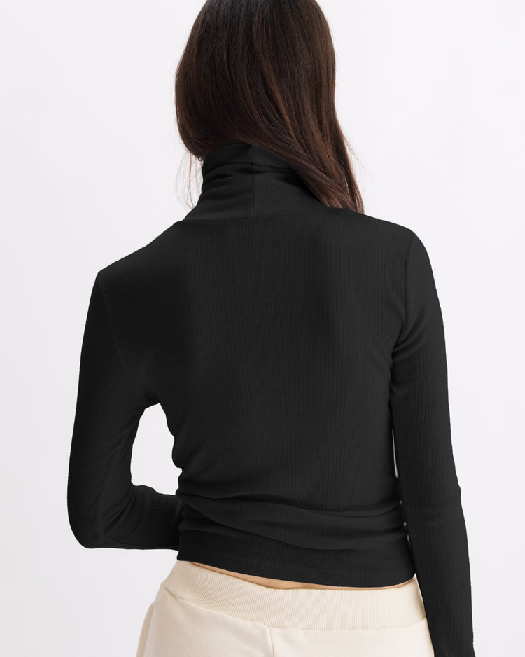 A woman with long brown hair, seen from behind, wears the Arlo Mott Clinton Long Sleeve Turtleneck in Black and light beige pants, standing against a plain white background.