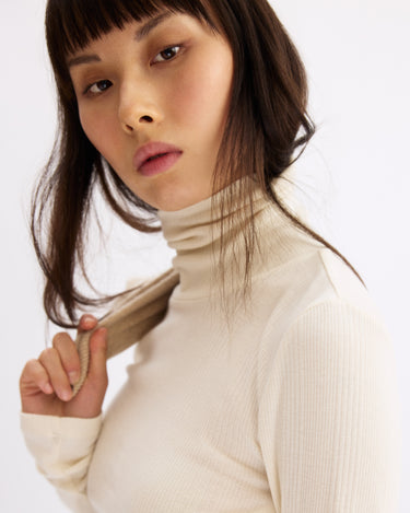 A woman with long brown hair and bangs confidently models the Arlo Mott Clinton Long Sleeve Turtleneck in Chalk, gently holding its collar against a plain white background.