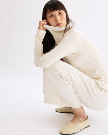 A woman with long dark hair squats on the floor in an off-white turtleneck top and Arlo Mott’s Kane Cropped Wide Sweatpants in Chalk, paired with beige slip-on shoes against a plain white background.