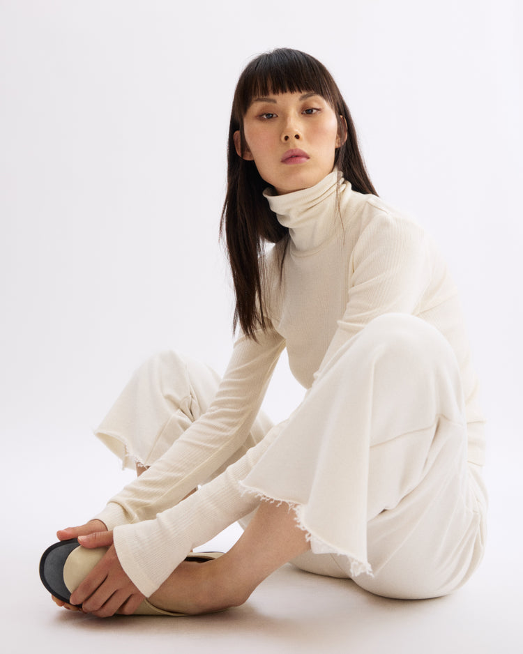 A woman with straight dark hair and bangs sits on the floor in a relaxed pose, wearing a cream turtleneck and Arlo Mott Kane Cropped Wide Sweatpants in Chalk, set against a plain white background.