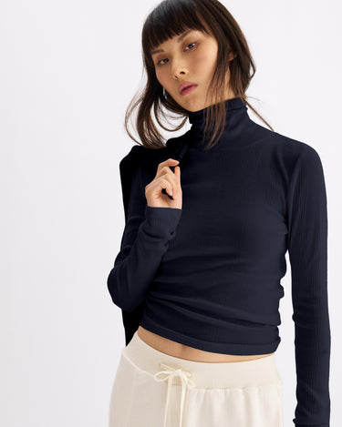 A woman with straight, dark hair and bangs models the Arlo Mott Clinton Long Sleeve Turtleneck in Navy, paired with cream drawstring pants, posing against a plain light background and holding her collar.
