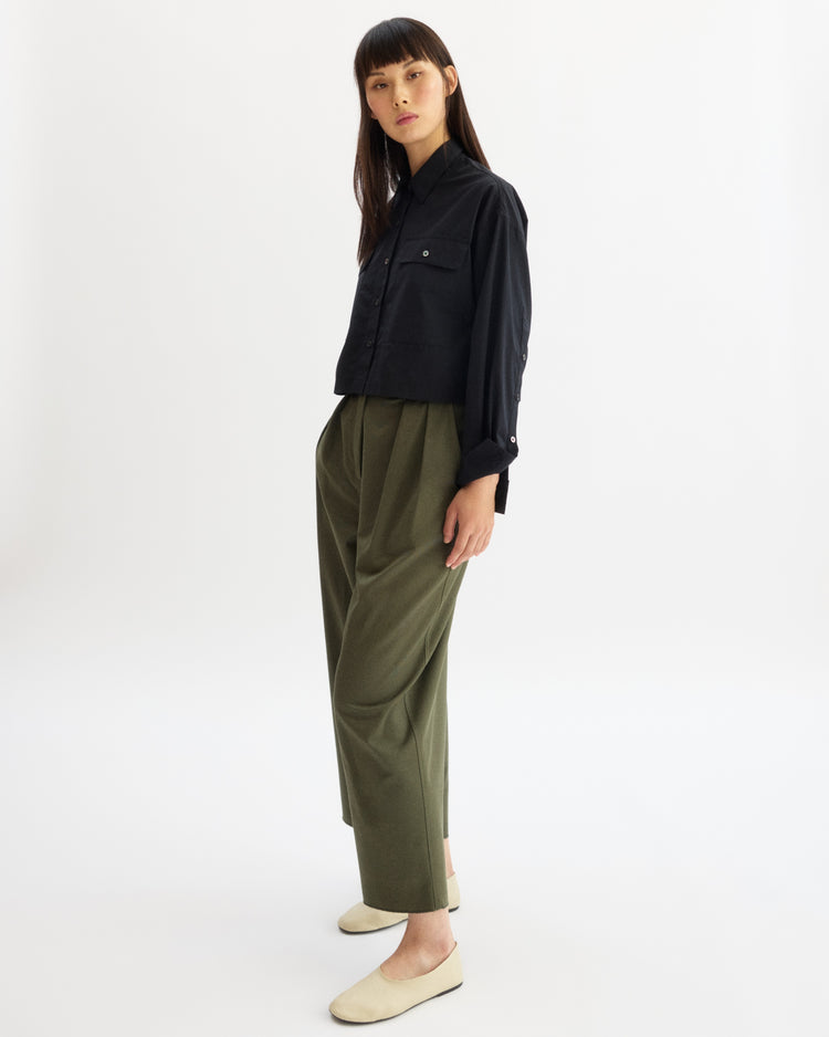 A woman stands in a minimal studio wearing the Arlo Mott Hester Boxy Short Button Down in Black, paired with loose olive green high-waisted trousers and beige flats. She has straight long hair and gazes slightly to the side against a white background.