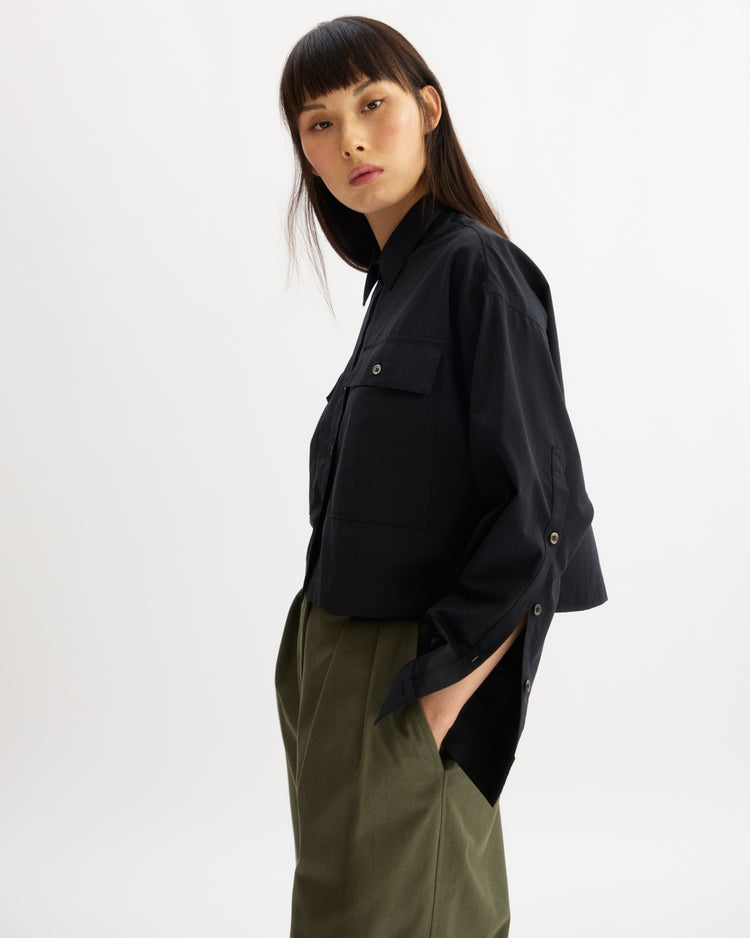 A woman with straight dark hair wears the Arlo Mott Hester Boxy Short Button Down in Black, styled loosely with large chest pockets and olive green high-waisted trousers, standing with hands in pockets against a plain light background.