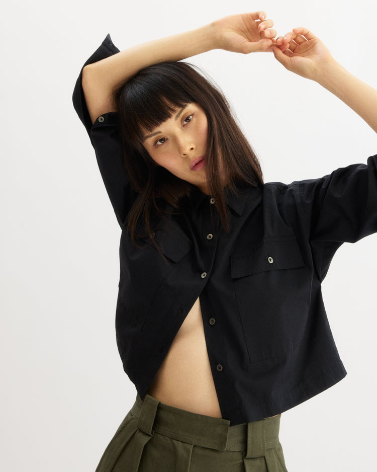 A person poses with arms raised and head tilted, wearing the Arlo Mott Hester Boxy Short Button Down in Black and high-waisted olive green pants against a plain white background.