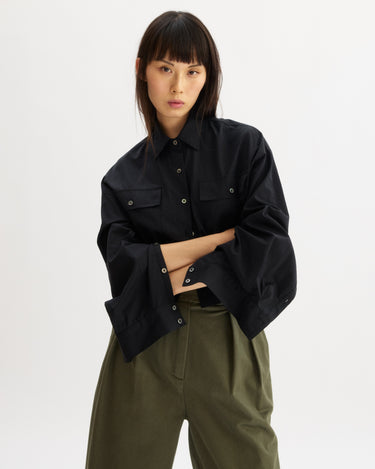 A woman with straight dark hair and bangs wears the Arlo Mott Hester Boxy Short Button Down in Black, styled with olive high-waisted pants, standing with arms crossed against a plain white background.