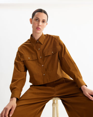 A person with short dark hair sits on a stool against a plain white background, wearing the Arlo Mott Hester Boxy Short Button Down in Cinnamon and matching brown pants.