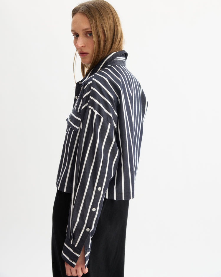 A woman with straight, light brown hair wears the Arlo Mott Hester Boxy Short Button Down in Navy Stripe, paired with a black skirt, as she poses against a plain white background.