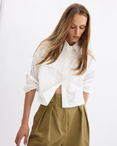 A woman with long blonde hair wears the Arlo Mott Hester Boxy Short Button Down in White and high-waisted khaki pants, looking down with her hand by her side against a plain white background.