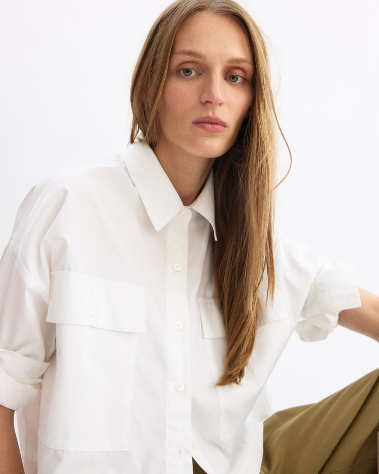 A woman with long, straight blonde hair wears the Arlo Mott Hester Boxy Short Button Down in White and looks at the camera with a neutral expression against a plain, light background.