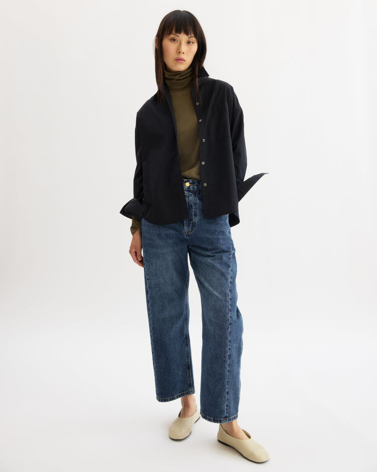 A woman with straight dark hair and bangs stands against a white background, wearing the Arlo Mott Watts Side Button Shirt in Black over an olive turtleneck, with blue wide-leg jeans and beige slip-on shoes.