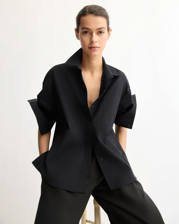 A person with dark hair pulled back wears the Arlo Mott Watts Side Button Shirt in Black and black pants, sitting on a stool against a plain white background.
