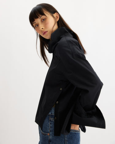 A woman with straight dark hair and bangs poses sideways in the Arlo Mott Watts Side Button Shirt in Black, styled with the collar up and paired with blue jeans against a plain white background.