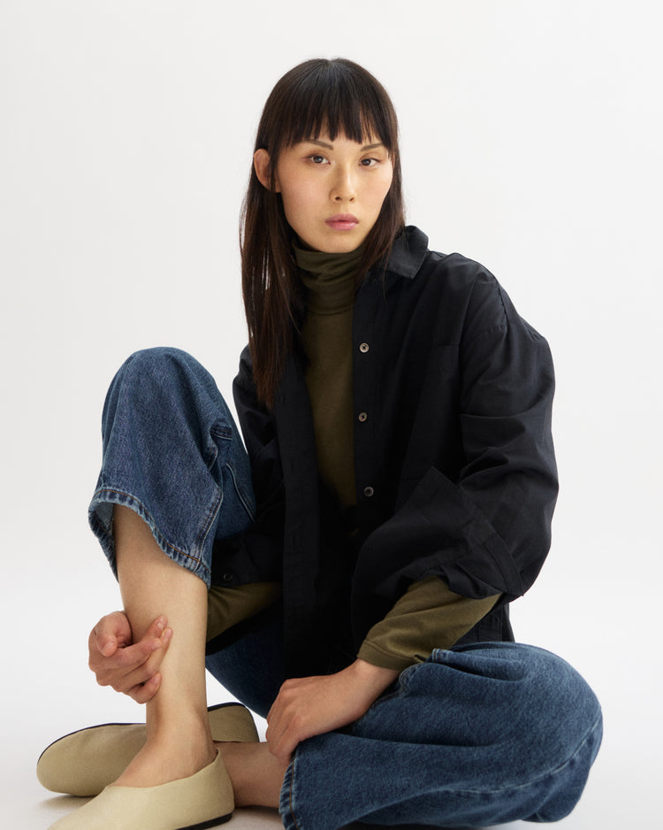 A person with straight dark hair and bangs sits against a white background, wearing the Arlo Mott Watts Side Button Shirt in Black over an olive turtleneck, blue jeans, and beige slip-on shoes, looking calmly at the camera.