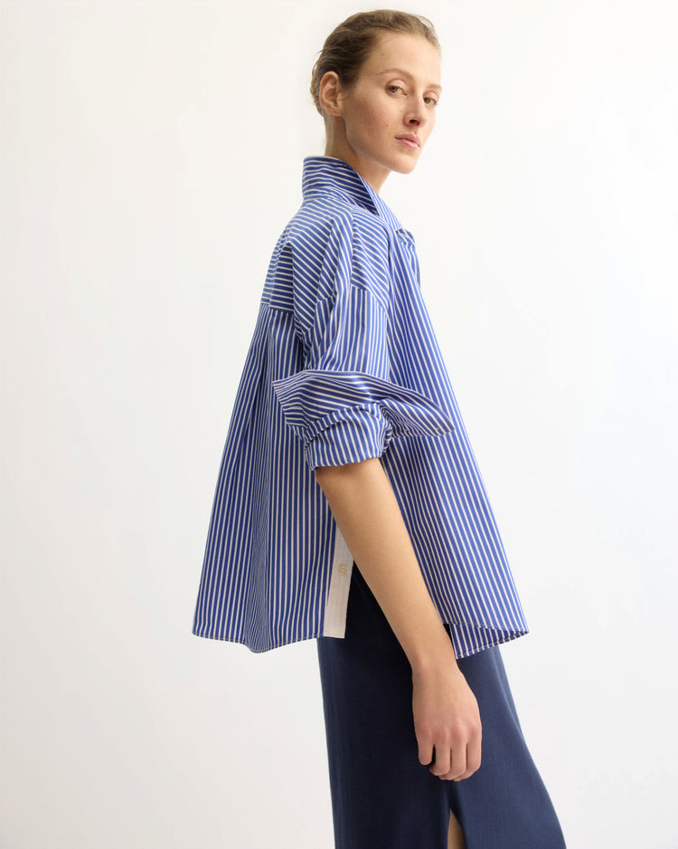 A person with light hair pulled back wears the Arlo Mott Watts Side Button Shirt in Blue Stripe, its sleeves rolled up, paired with a navy slit skirt, standing against a plain light background.