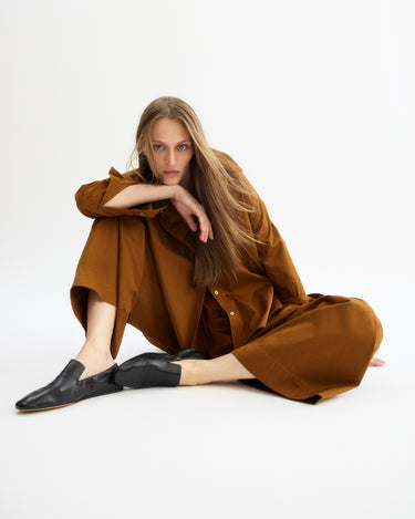 A woman with long, straight hair sits on the floor in a relaxed pose, wearing Arlo Mott’s Hudson Pull-on Pants in Cinnamon and black shoes, set against a plain white background.
