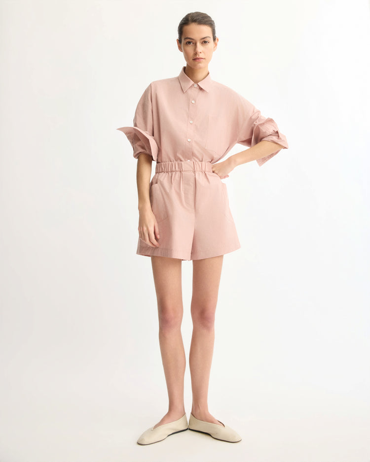 A woman stands against a white background wearing the Arlo Mott Watts Side Button Shirt in Dusty Rose with rolled sleeves, matching elastic-waist shorts, and cream flats.